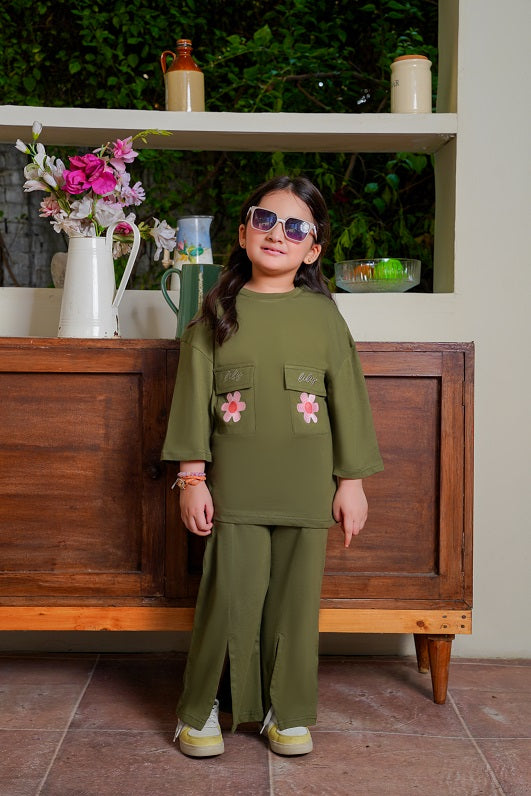 Child wearing an olive green outfit with floral details, standing in front of a wooden cabinet.