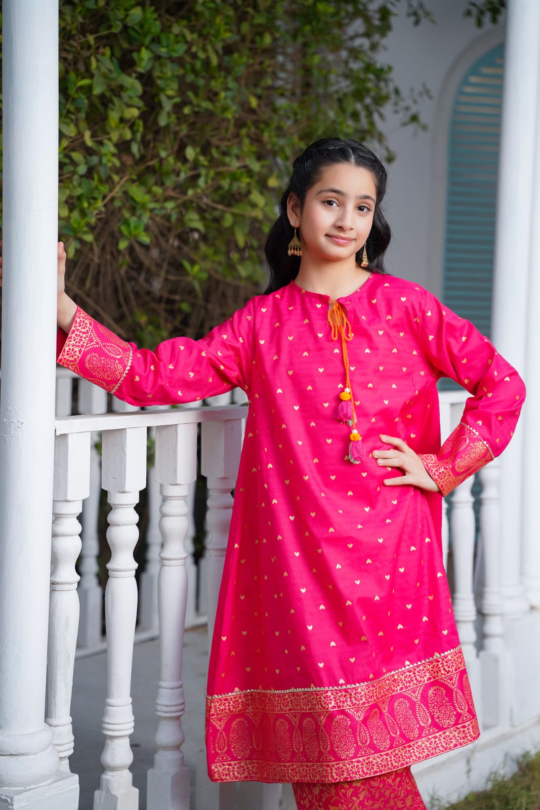 Young girl wearing a bright pink traditional outfit with white patterns, standing on a white railing.