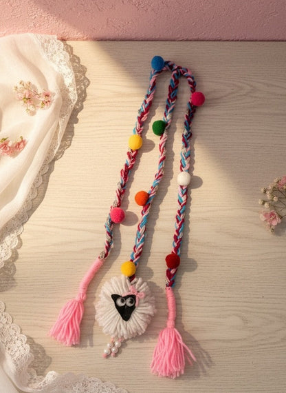Lily braided yarn hair accessory with colorful pom-poms, pink tassels, and a sheep charm with googly eyes and pearl accents.