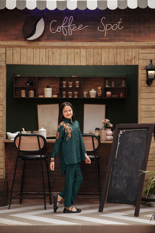 Lily girl wearing an emerald green button-down shirt and trousers with golden floral embroidery in a cafe setting