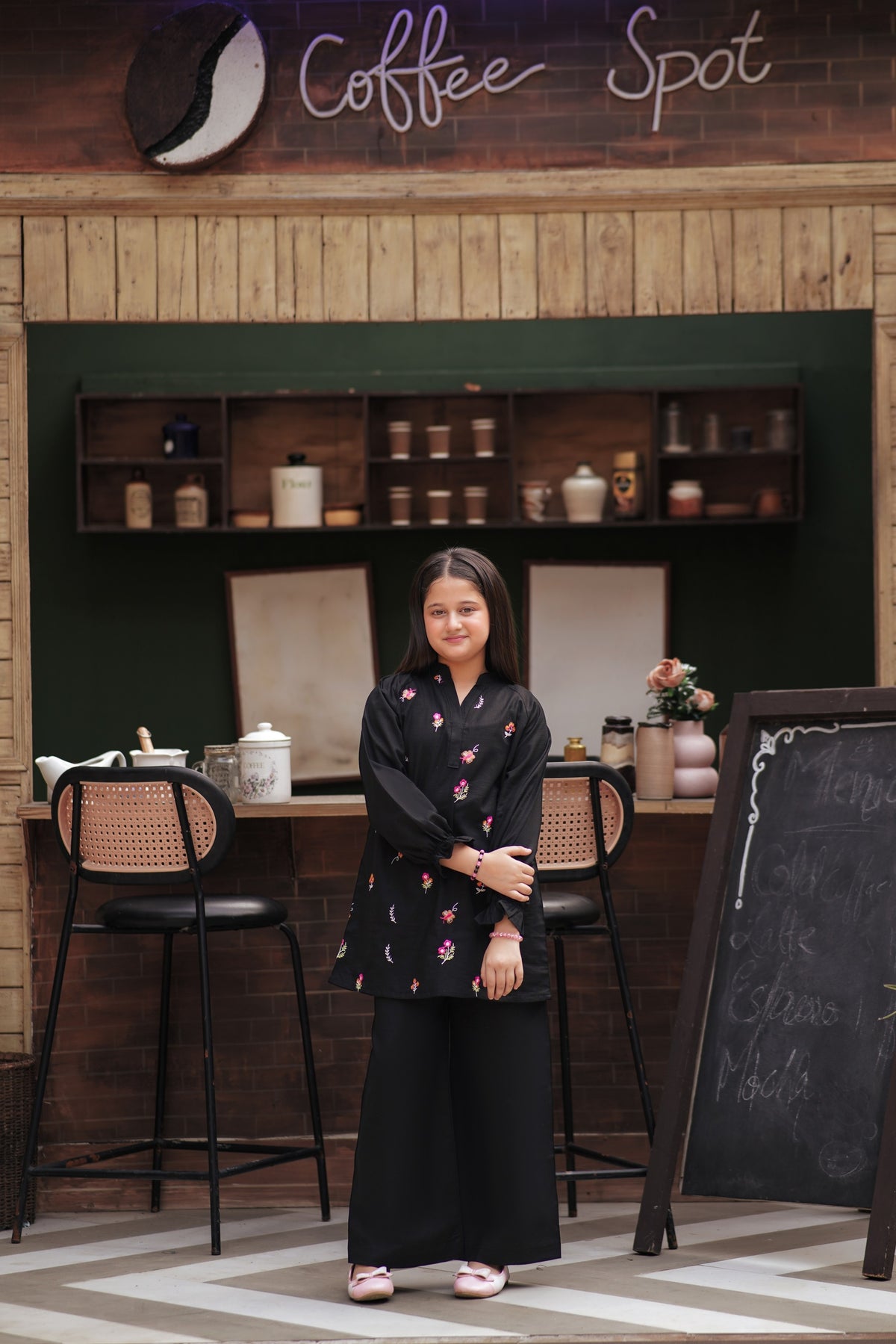 Lily girls black tunic and trouser set with multi-colored floral embroidery, V-neck, and ruffled cuffs in a cafe