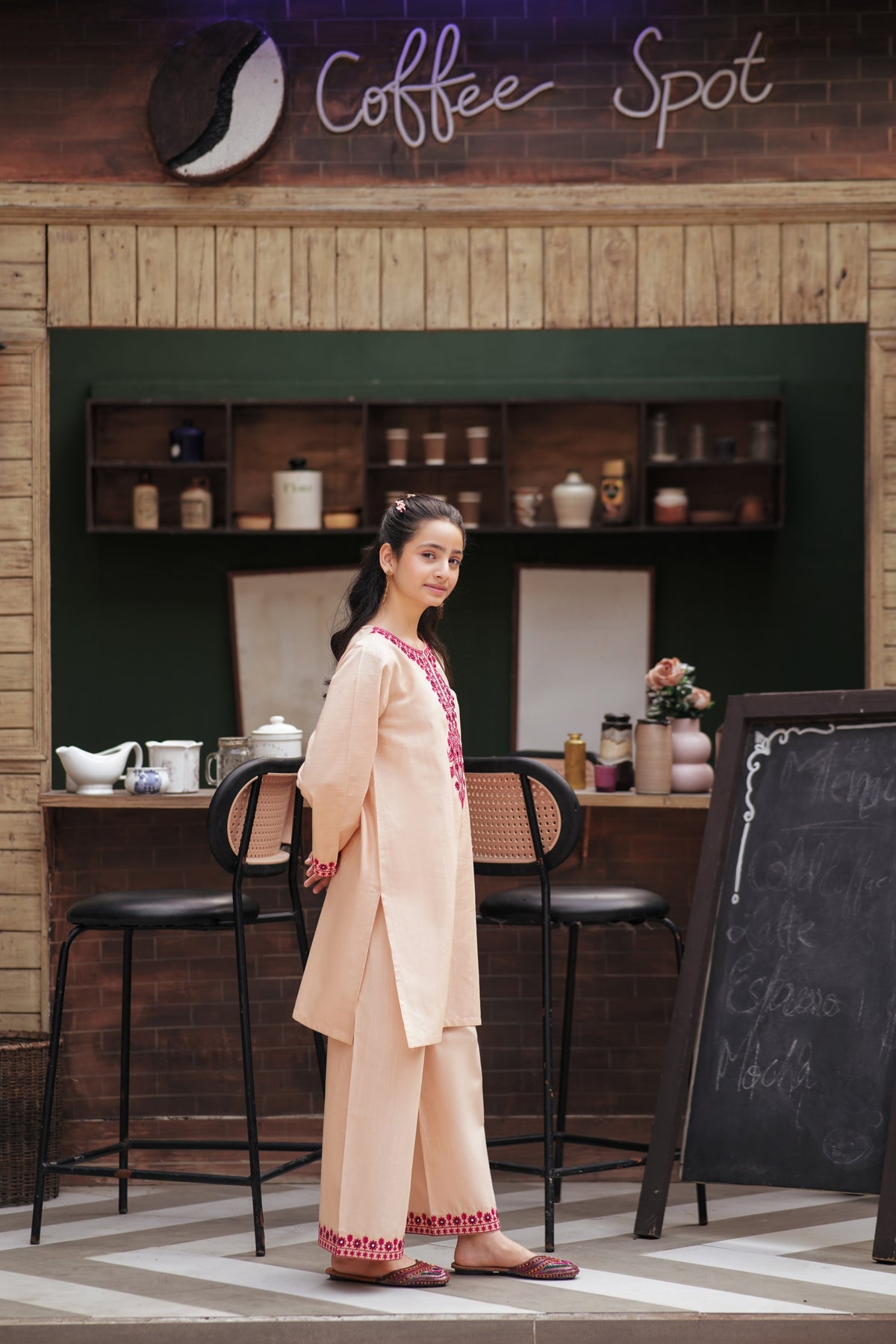 Lily girls peach kurta and trouser set with magenta floral embroidery and mirror work, worn by a girl in a cafe.
