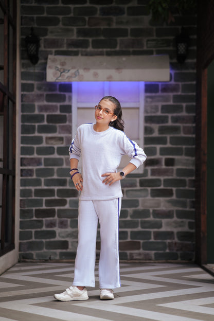 Lily girls white two-piece tracksuit with purple side stripes, worn by a girl with glasses against a brick wall.