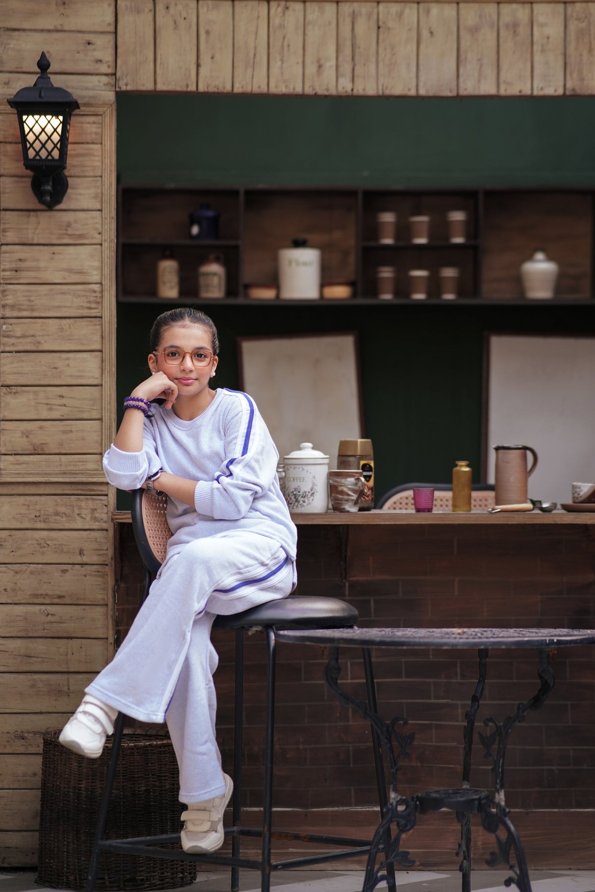 Lily girl wearing a white textured two-piece tracksuit with purple side stripes, sitting on a stool in a cafe setting.