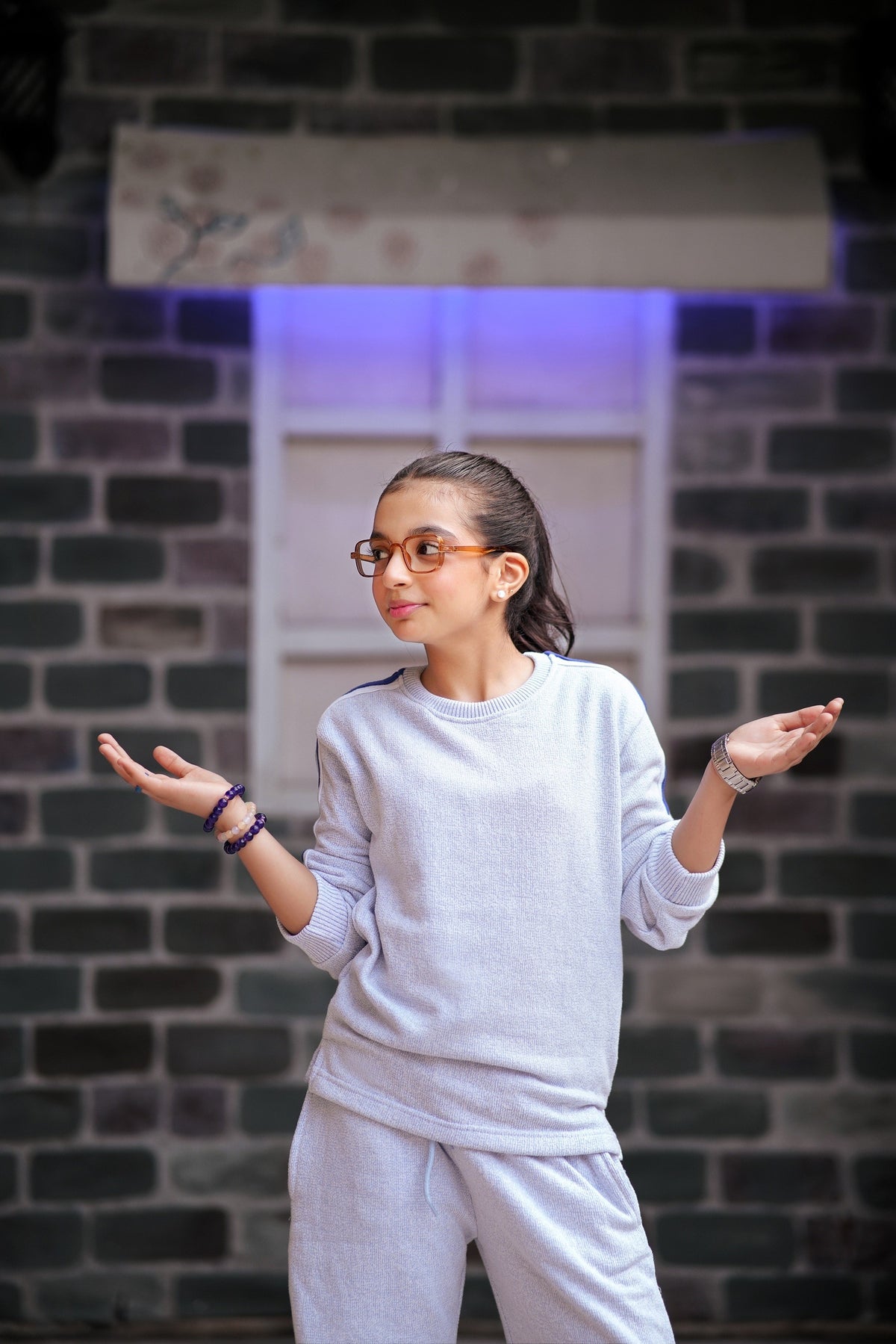 Lily girls white textured two-piece tracksuit with purple side stripes worn by a girl in glasses against a brick wall
