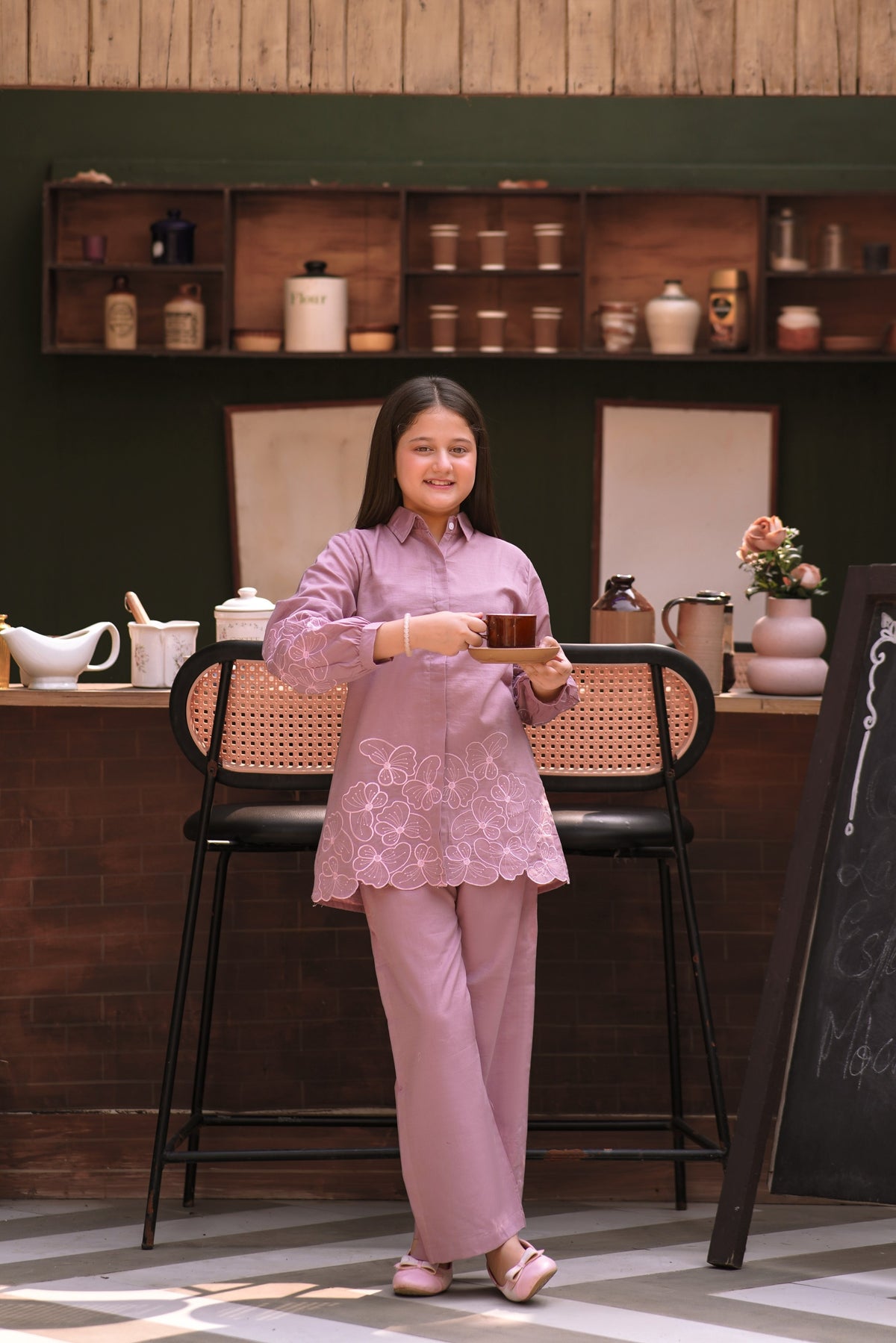 Lily girl wearing a mauve button-down shirt and trousers set with floral embroidery, holding a cup in a cafe setting.