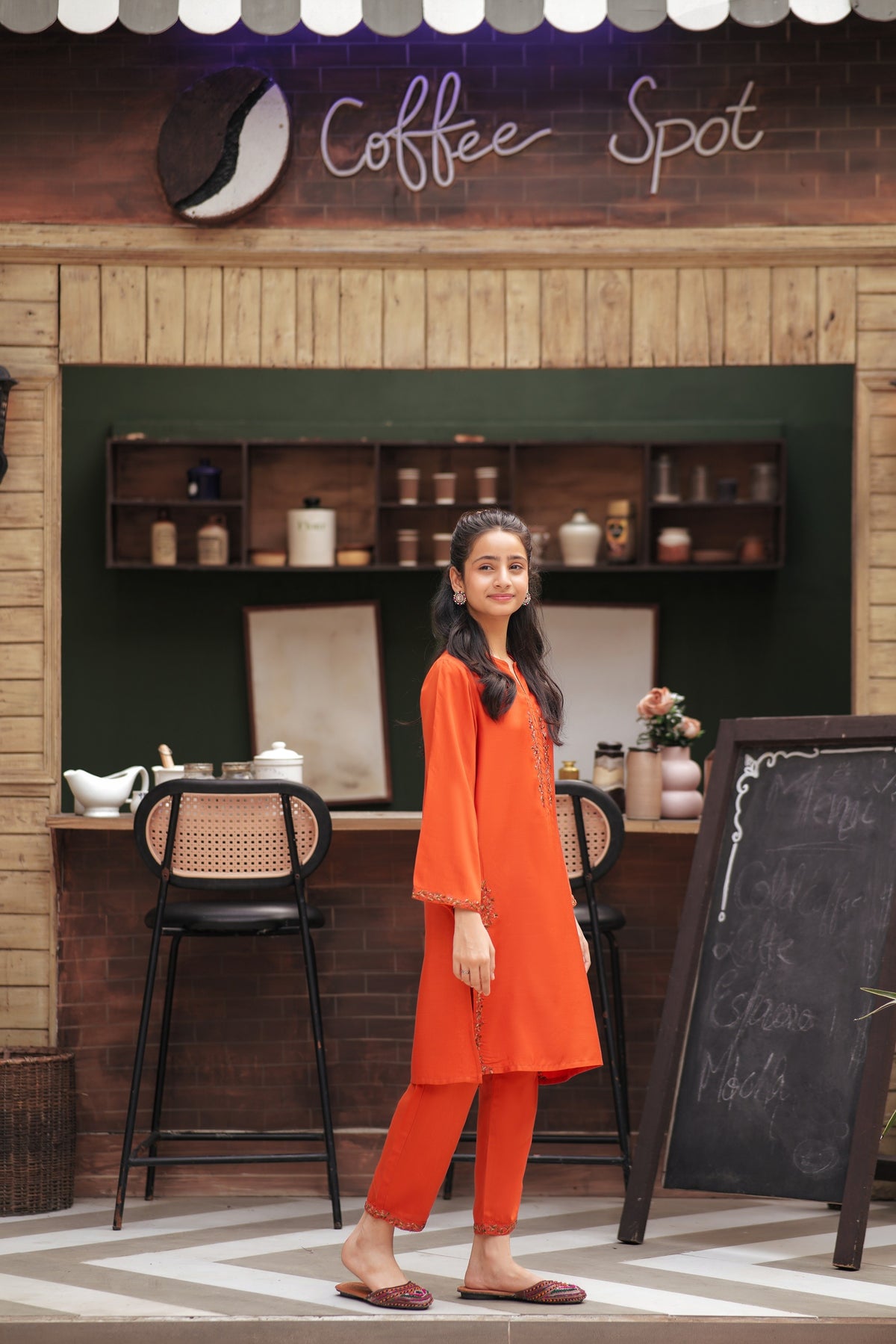 Lily girl wearing a vibrant orange embroidered kurta and trouser set with floral details in a cafe setting
